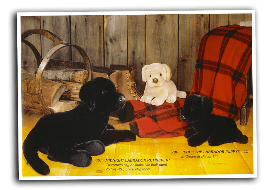 A plush Black Lab stuffed animal lies near a pair of all-weather boots and a pile of firewood. Two plush Labrador puppies sit nearby on a wool buffalo plaid blanket