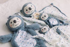 All four pieces from the DOUGLAS Baby Indy Otter character collection are laid out on display. The collection contains the Blanki Lovey, Lil' Snuggler Teether toy, Paci Lovey, and the soft, plush Sshlumpie.