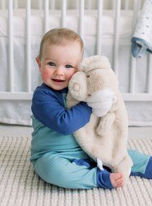 A cheerful baby boy dressed in a blue romper smiles at the camera as he snuggles a plush Sshlumpie dog from the DOUGLAS Baby Auggie Tan Puppy character collection up against the side of his face.
